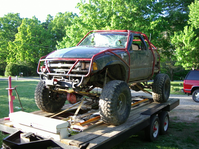 Off-road truck with roll cage and large tires on a trailer, red SUV in background