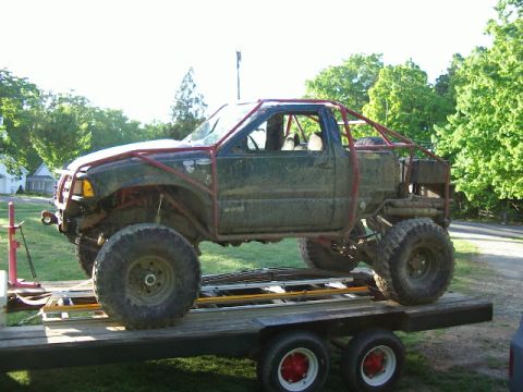 1985 Ford Bronco - A heavily modified pickup truck with a custom roll cage and oversized tires is loaded onto a trailer.