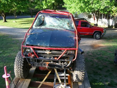 1985 Ford Bronco - A heavily modified off-road truck with a damaged windshield is loaded onto a trailer, with a red pickup truck in the background.