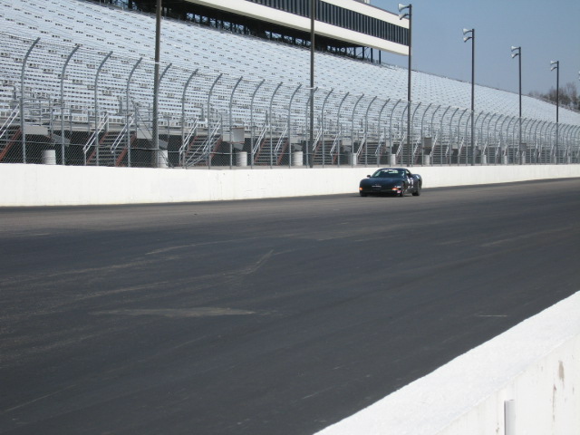 Black coupe on a race track, front view, with grandstands in the background