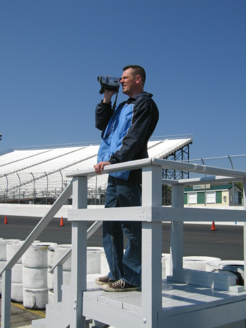 1991 Ford LTD Crown Victoria - A man films with a camcorder from a raised platform at a racetrack, with empty grandstands visible under a clear blue sky.