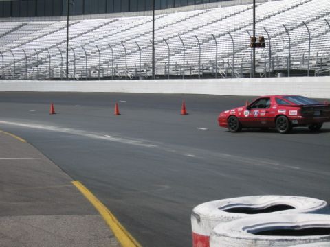 1991 Ford LTD Crown Victoria - A red sports car navigates a race track with cones and grandstands visible.