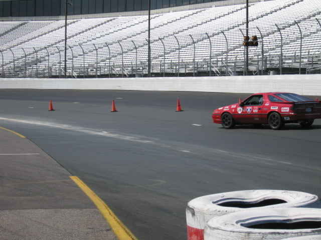 Red sports car with racing decals on a track, cones in foreground, grandstands in background