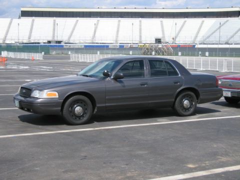1991 Ford LTD Crown Victoria - A dark gray sedan is parked in an asphalt lot with stadium seating visible in the background.