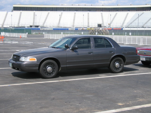 Dark gray sedan parked in a lot with stadium seating in the background. Another car is partially visible to the right