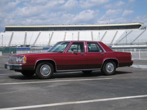 1991 Ford LTD Crown Victoria - A red sedan is parked in a lot with a large stadium in the background.