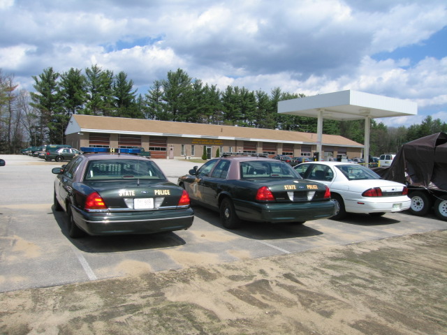 Two dark green police cars with light bars are parked, one behind the other. A white sedan is parked to the right