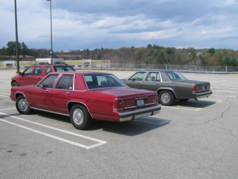 1991 Ford LTD Crown Victoria - Two sedans, a red one and a gray one, are parked in an outdoor lot under a cloudy sky.