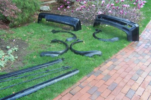 1996 Ford Bronco - Various black car body parts, including bumpers and fender flares, are arranged on a lawn next to a brick pathway.
