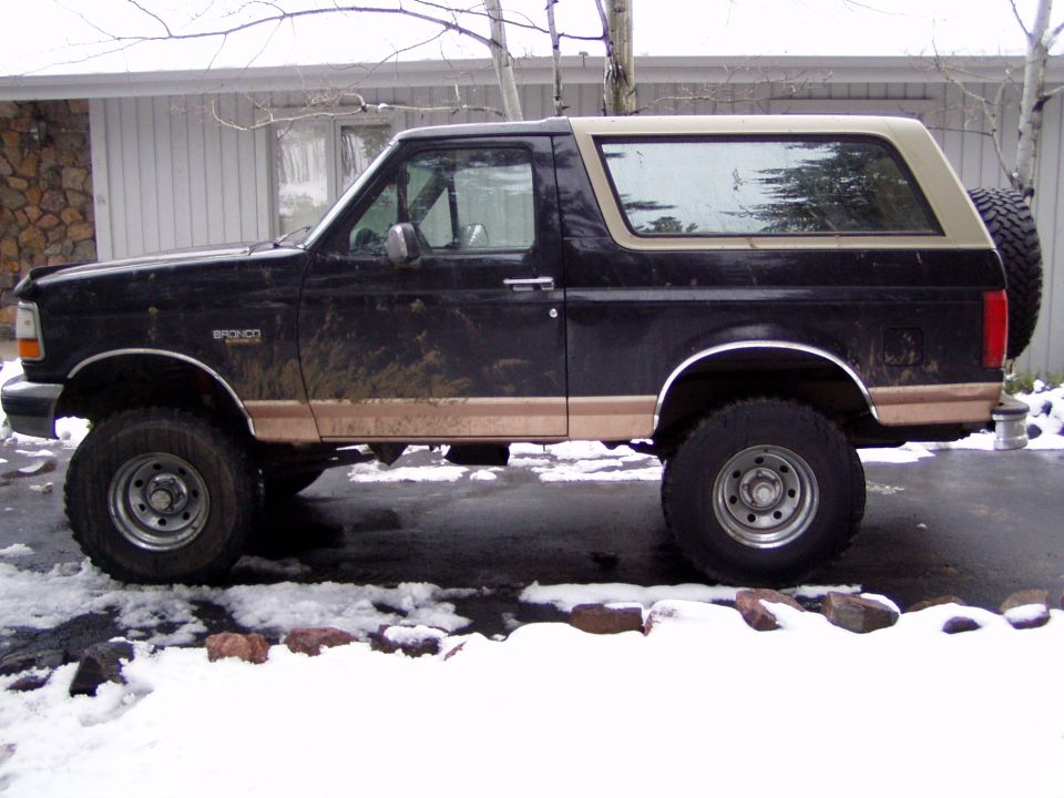 Muddy black and tan lifted Ford Bronco side view on snowy driveway with rear-mounted spare tire