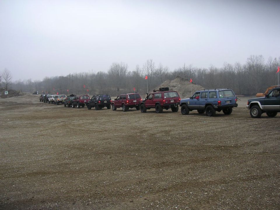Line of Jeep Cherokees parked on a dirt lot, some with roof racks. Trees and overcast sky in background