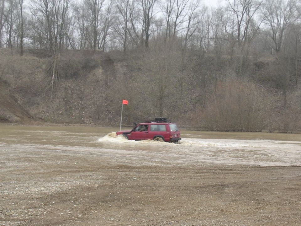 Red Jeep Cherokee driving through a shallow body of water with a flag on a pole nearby