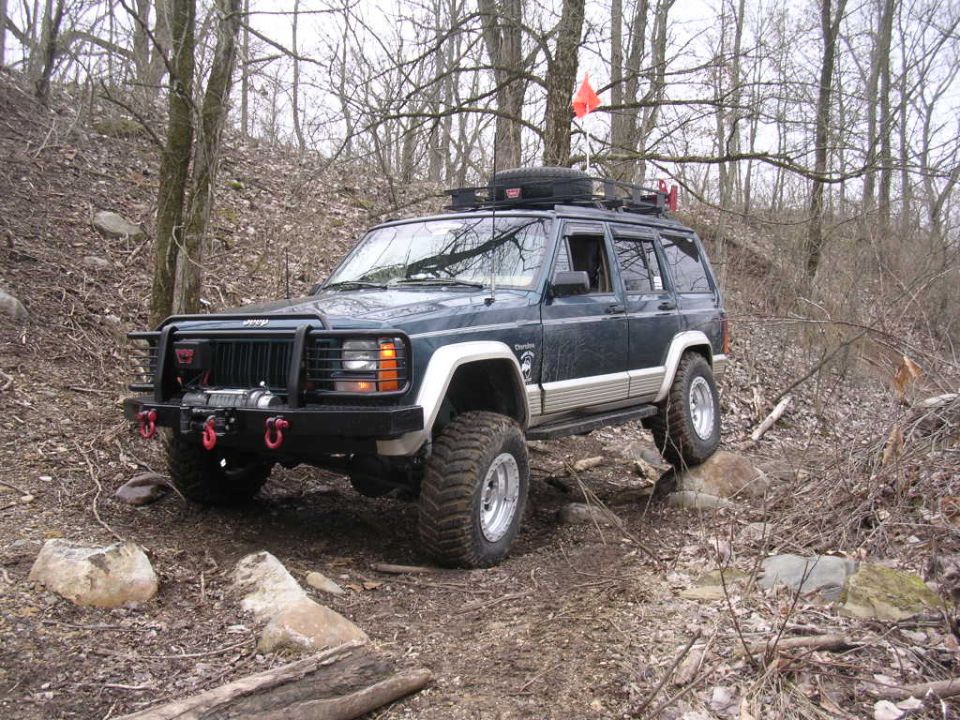 Dark green Jeep Cherokee on a rocky, wooded trail. It has a winch, roof rack, and an orange flag on top