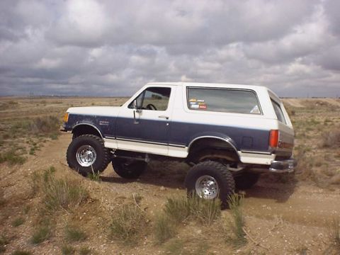1988 Ford Bronco - This lifted Ford Bronco is parked on a dirt mound under a cloudy sky.