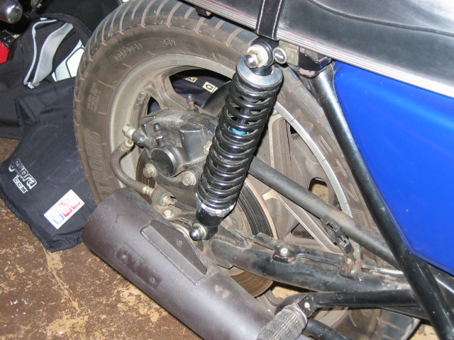 motorcycle rear wheel, black shock absorber, and exhaust pipe. Black bags with 'Suara.com' text are on the left
