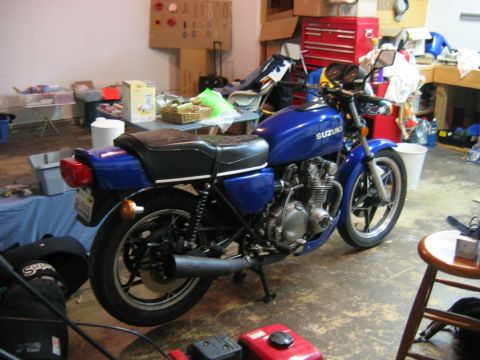 1985 Ford LTD Crown Victoria - A blue Suzuki motorcycle is parked in a cluttered garage, with tools and storage items visible in the background.