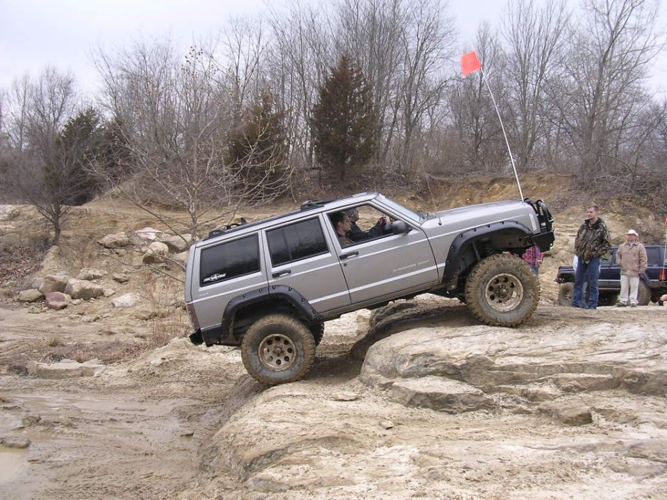 Silver Jeep Cherokee climbing over rocks with an orange flag on antenna. Another Jeep and a pickup truck are visible in the background