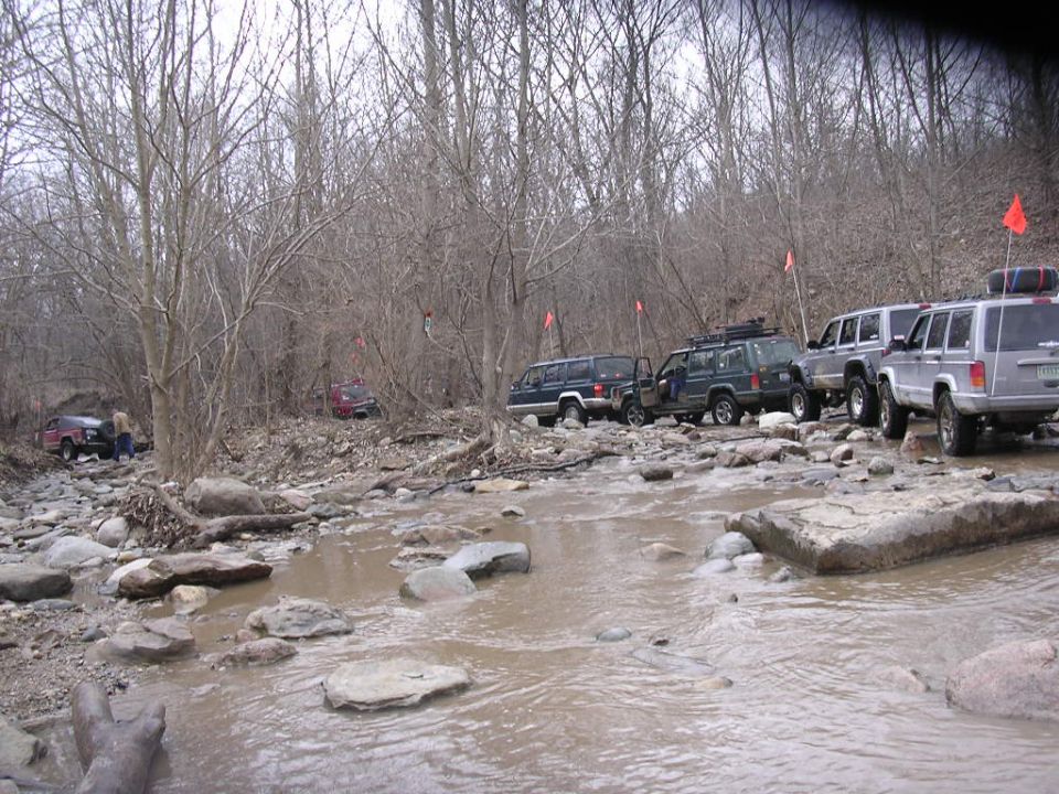Several Jeep Cherokees driving through a rocky, muddy stream in a wooded area. Some have roof racks and flags