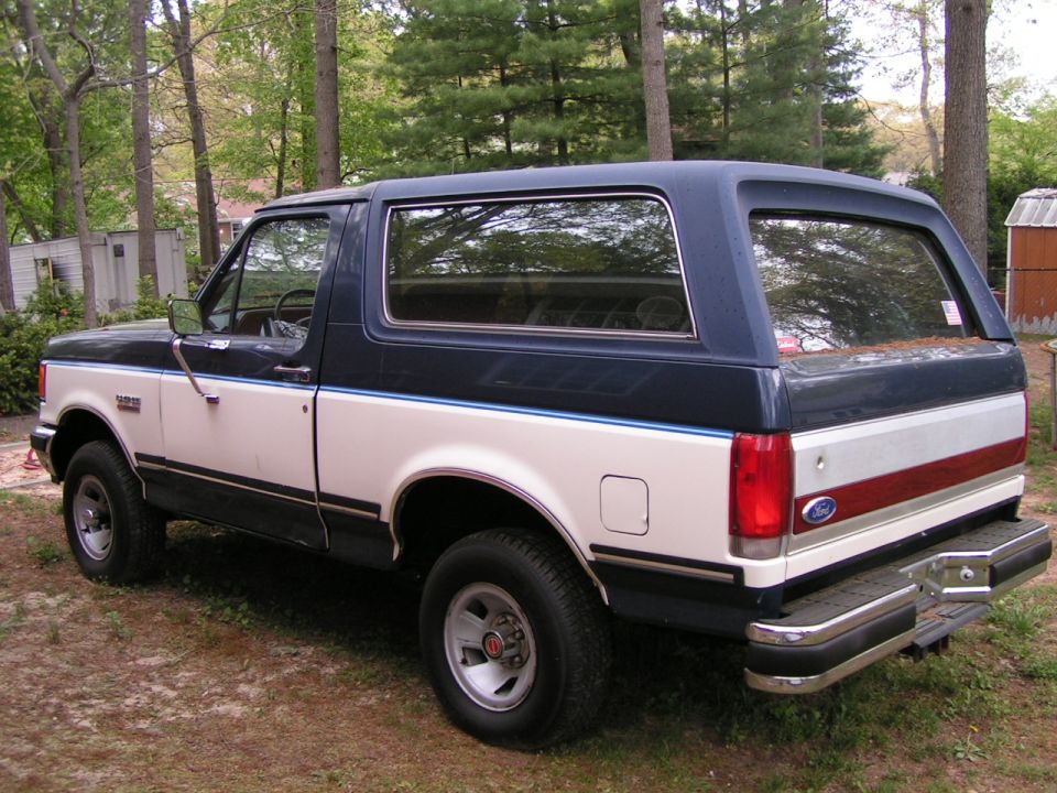 Two-tone blue and white Ford Bronco rear 3/4 view parked on grass under pine trees