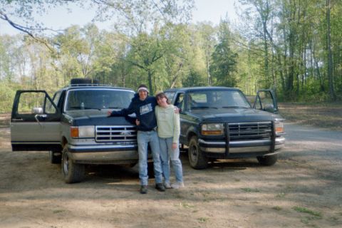 1988 Ford Bronco - Two Ford Broncos are parked outdoors with trees in the background. The vehicle on the left has its driver's door open.