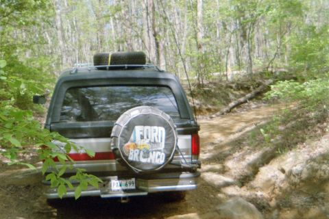 1988 Ford Bronco - A black Ford Bronco navigates a wooded dirt path, its spare tire cover clearly displaying 'Ford Bronco'.