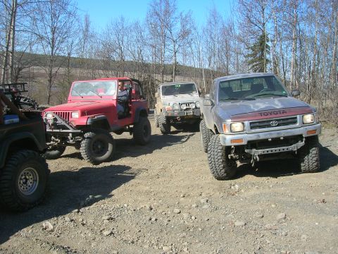 2003 Jeep TJ - Several off-road vehicles, including a red Jeep Wrangler and a Toyota pickup, are parked on a gravelly surface surrounded by trees.