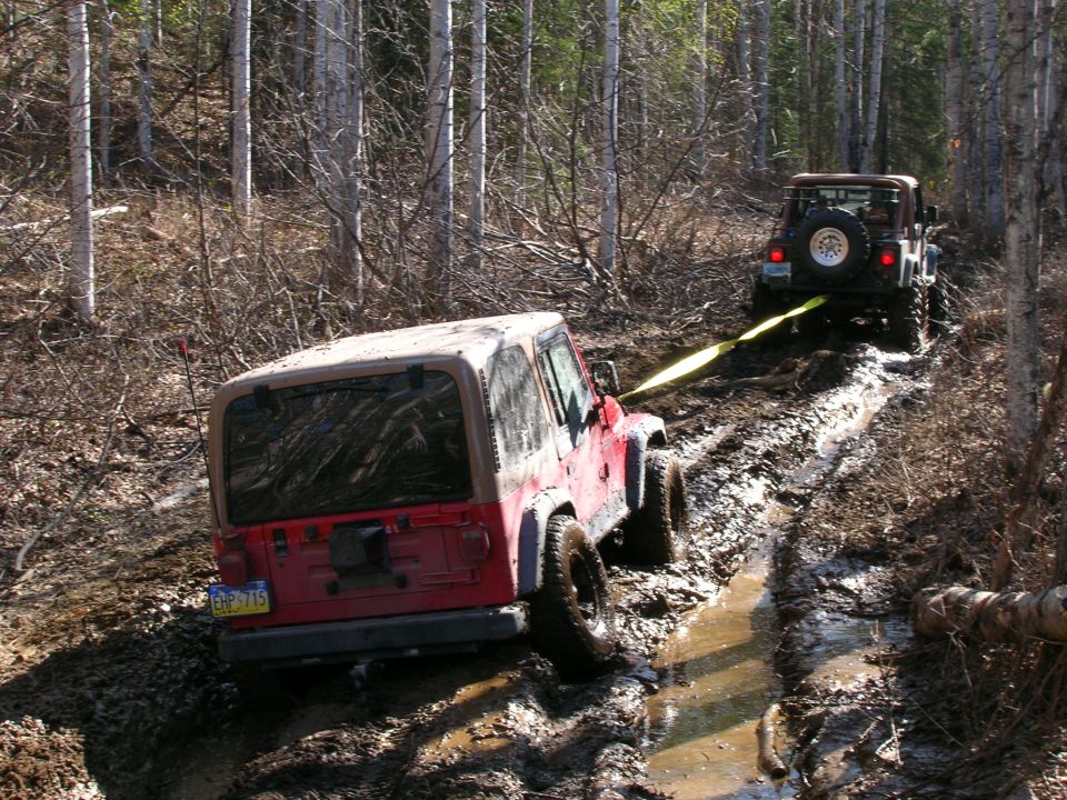 Red Jeep TJ being pulled out of mud by another Jeep in a wooded area