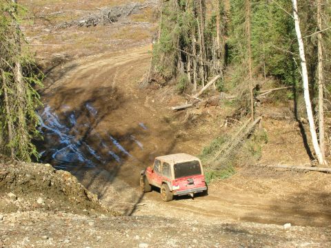 2003 Jeep TJ - A red Jeep Wrangler navigates a muddy, off-road trail surrounded by trees.