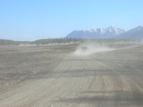 2003 Jeep TJ - A vehicle drives down a dusty dirt road, leaving a trail of dust in its wake, with mountains visible in the distance.