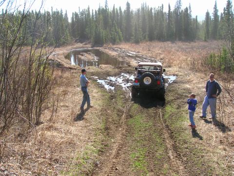 2003 Jeep TJ - A Jeep Wrangler navigates a muddy off-road trail surrounded by trees and dry brush.
