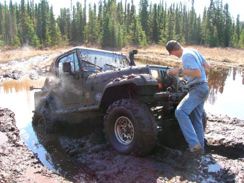 2003 Jeep TJ - A 2003 Jeep TJ with a snorkel is stuck in deep mud while a person inspects the engine.