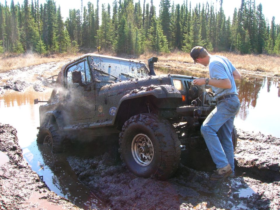 Mud-covered 2003 Jeep TJ with snorkel, stuck in a muddy puddle with a person checking the engine