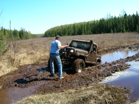 2003 Jeep TJ - A Jeep is stuck in a muddy trail surrounded by trees and dry grass.