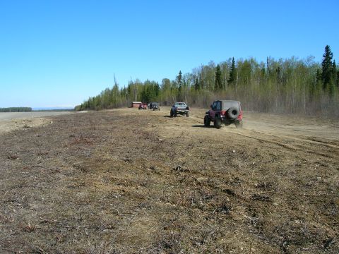 2003 Jeep TJ - Several off-road vehicles drive on a dirt path near a wooded area under a clear blue sky.