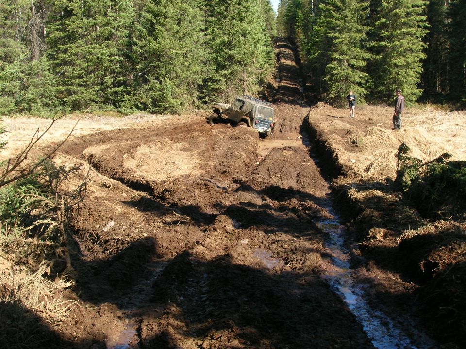Jeep stuck in a muddy ditch on a wooded trail, two people watching