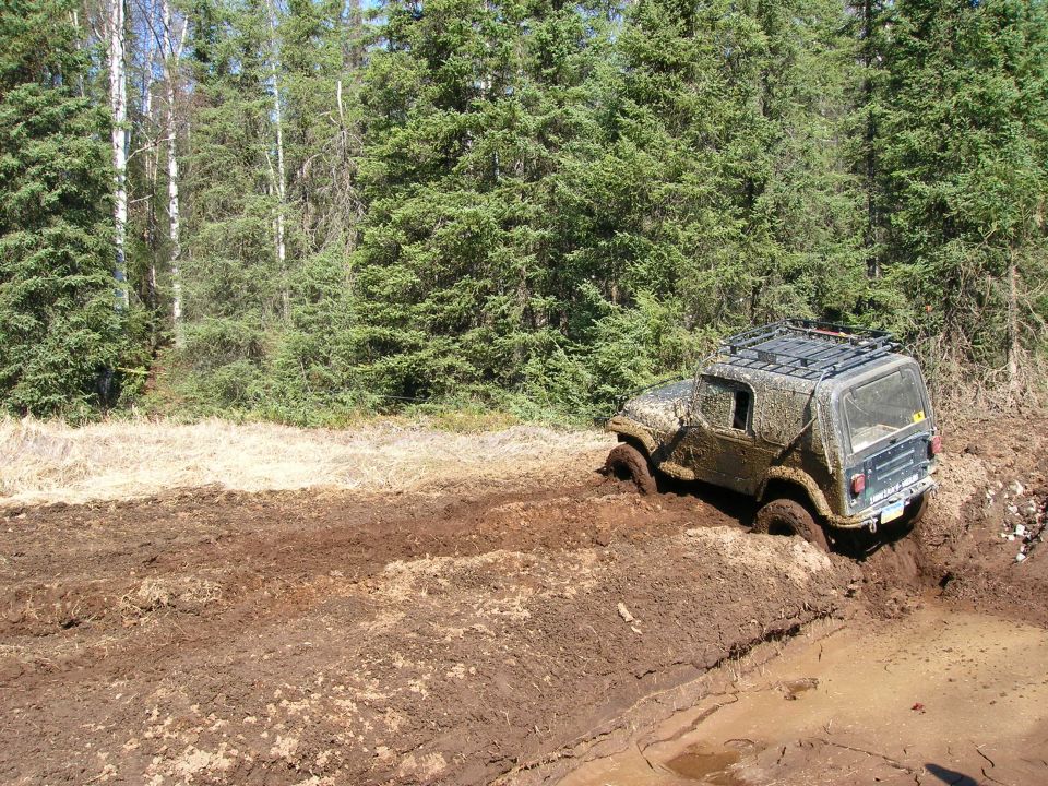 Mud-covered 2003 Jeep TJ stuck in a muddy rut on a wooded trail