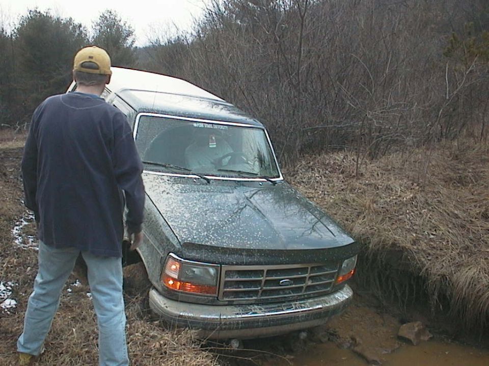 Man in yellow hat watches a 1992 Ford Bronco stuck in a muddy ditch