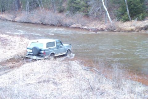 1992 Ford Bronco - A Ford Bronco navigates a river crossing surrounded by a wooded area.