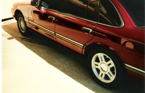 1992 Ford Crown Victoria - Angled driver-side view of a dark red early-1990s Ford Crown Victoria sedan in bright sunlight, from a scanned film print. The car wears mi…