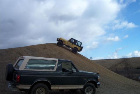 1992 Ford Bronco - A dark green Ford Bronco is parked while a yellow Jeep Wrangler ascends a dirt incline under a cloudy sky.