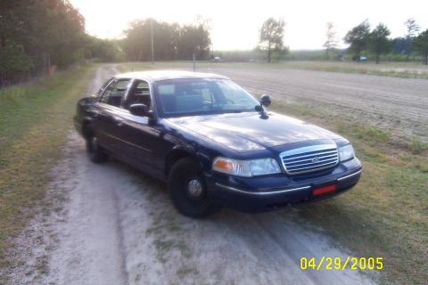 1998 Ford Crown Victoria - A dark blue Ford Crown Victoria Police Interceptor is parked on a dirt road with fields in the background.