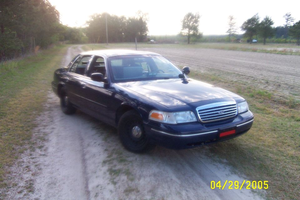 Dark blue Ford Crown Victoria sedan on dirt road, front three-quarter view, black steel wheels