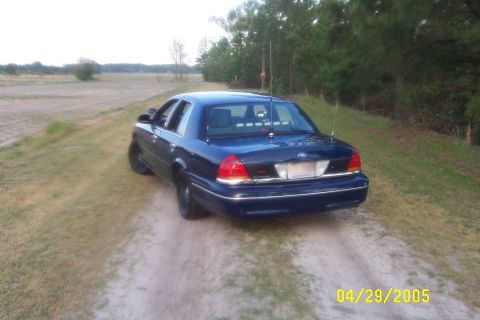 1998 Ford Crown Victoria - A dark blue 1998 Ford Crown Victoria is seen from the rear on a dirt road in a rural setting.
