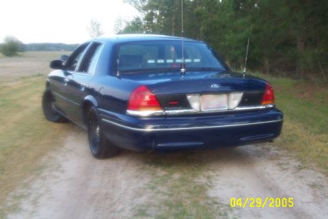 1998 Ford Crown Victoria - Rear view of a dark blue 1998 Ford Crown Victoria on a dirt road with trees in the background.