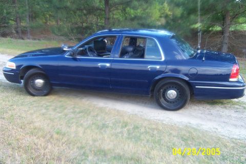 1998 Ford Crown Victoria - Driver-side profile of a dark blue Ford Crown Victoria four-door sedan parked on a grassy dirt lane in front of pine trees. It wears black…