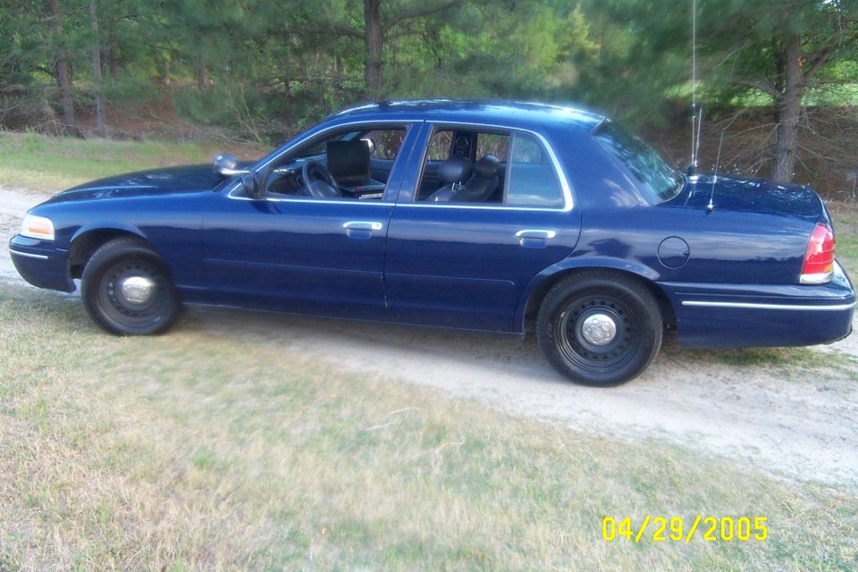 Dark blue Ford Crown Victoria sedan side view on dirt lane with black steel wheels