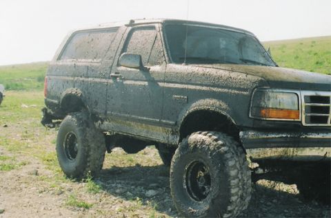 1992 Ford Bronco - Passenger-side three-quarter front view of a lifted Ford Bronco caked in wet mud from roof to rockers, riding on large mud-terrain tires wi…