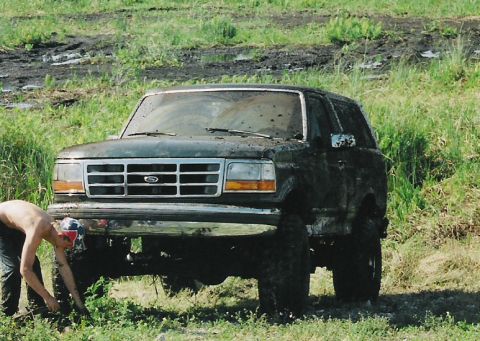 1992 Ford Bronco - A muddy 1992 Ford Bronco sits in a grassy field with a person attempting to free it from the mud.