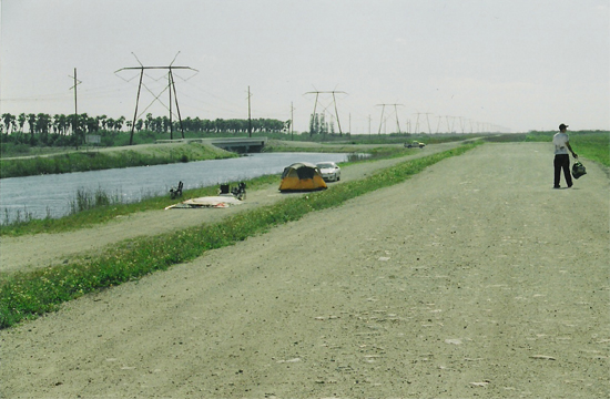 Power lines stretch across a landscape with a canal and a dirt road where a person stands