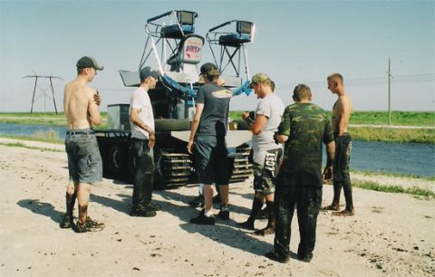 1992 Ford Bronco - Seven mud-splattered men stand around a blue-and-white tracked swamp buggy parked on a dirt levee beside a canal, its tall rear tower carry…
